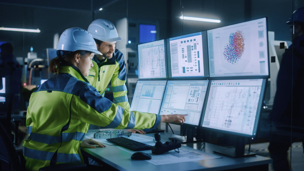 Two employees in hardhats and hi-vis jackets discussing a project by a computer screen.